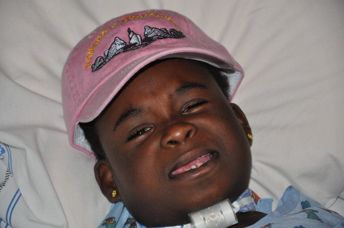 Child smiling, wearing pink hat in bed.