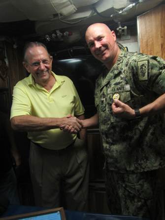 Two men shaking hands in a casual indoor setting, one in a yellow shirt and the other in camouflage.